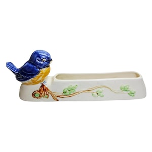 May include: A ceramic planter with a bluebird design. The bird is blue and yellow, perched on a brown branch with green leaves. The planter has a cream-coloured rectangular base.