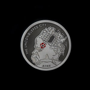 Könnte beinhalten: Eine silberne Münze mit einem Schwarzweißbild eines Personenkopfes mit einem Gebäude darauf. Der Text "THE UNCONQUERED CITY" ist um den Rand der Münze herum geschrieben. Das Jahr "2022" ist am unteren Rand der Münze geschrieben.