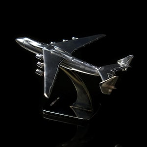 May include: A silver model airplane of a large cargo plane with six engines. The plane is on a black stand.
