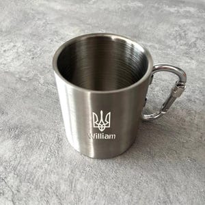 Pode incluir: Uma caneca de aço inoxidável prateada com uma alça de mosquetão. A caneca tem um símbolo de tridente e o nome "William" gravado no lado.