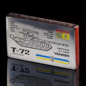 Può includere: Un oggetto metallico rettangolare con un'illustrazione dettagliata di un carro armato T-72, con specifiche tecniche e la scritta "LET THEM VANISH". L'oggetto ha un bordo rosso e un simbolo di pericolo.