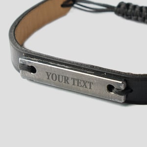Könnte beinhalten: Schwarzes Lederarmband mit einer silbernen Metallplatte, die mit dem Text "YOUR TEXT" graviert ist.