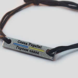 Könnte beinhalten: Schwarzes Kordelarmband mit rechteckiger Metallplatte. Die Platte trägt den ukrainischen Text "Слава Україні Героям слава" mit blauen und gelben Streifen. Das Armband befindet sich auf weißem Hintergrund.