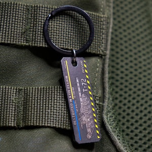 May include: A metal keychain with a black key ring and a silver tag. The tag features a black and white illustration of a tank with the text "DESTROYED IN KYIV REGION 24.02.2023" and "T-72 001/200".
