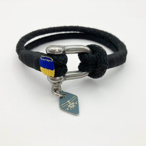 Könnte beinhalten: Schwarzes Seil-Armband mit einem silbernen Verschluss und einem kleinen blau-gelben Flaggen-Anhänger. Der Anhänger hat ein silbernes Helikopter-Design.