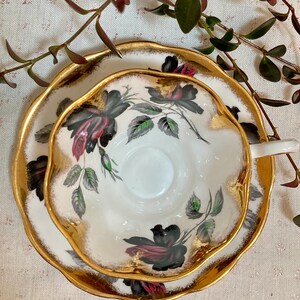 May include: A white teacup and saucer with a black rose pattern and gold trim. The cup and saucer have a scalloped edge.