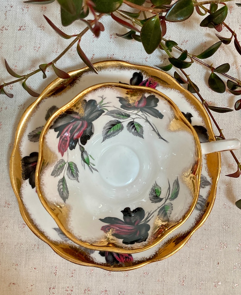 May include: A white teacup and saucer with a black rose pattern and gold trim. The cup and saucer have a scalloped edge.