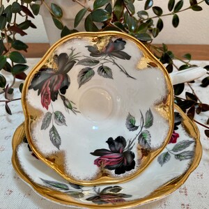 May include: A white teacup and saucer with a black and pink rose pattern and gold trim. The cup and saucer have a scalloped edge.