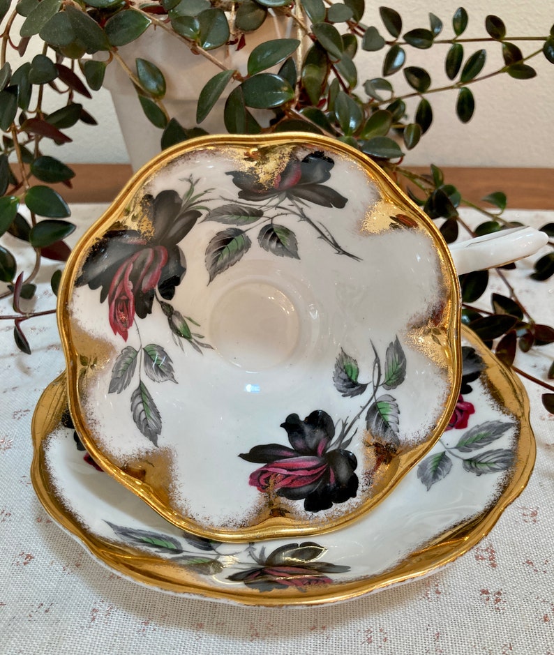 May include: A white teacup and saucer with a black and pink rose pattern and gold trim. The cup and saucer have a scalloped edge.