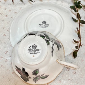 May include: A white teacup and saucer with a black and green floral design. The bottom of the cup and saucer have the words "Royal Albert Bone China England" printed on them.
