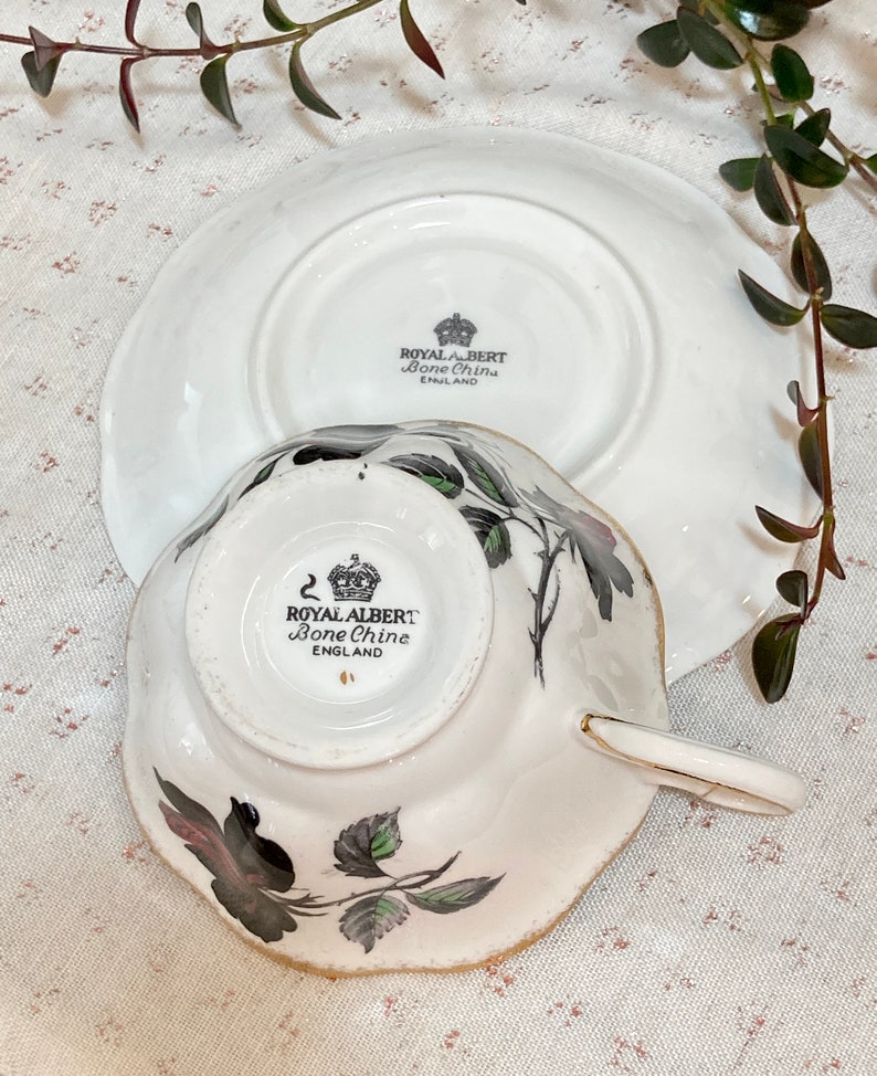 May include: A white teacup and saucer with a black and green floral design. The bottom of the cup and saucer have the words "Royal Albert Bone China England" printed on them.