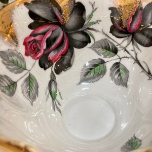 May include: A white teacup with a gold rim and black and red roses with green leaves. The teacup has a white interior.