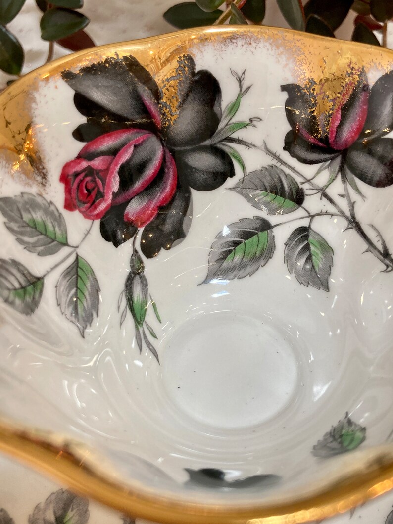 May include: A white teacup with a gold rim and black and red roses with green leaves. The teacup has a white interior.