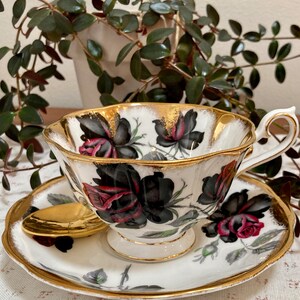 May include: A white teacup and saucer with a black and red rose pattern and gold trim. The cup and saucer are on a white tablecloth.