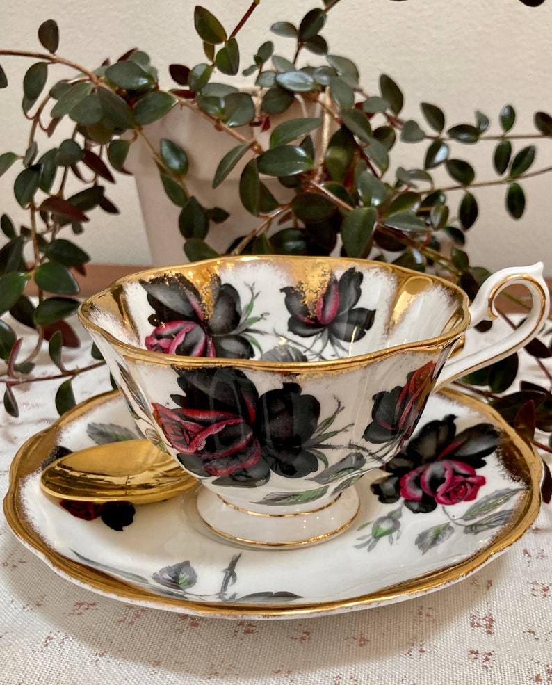 May include: A white teacup and saucer with a black and red rose pattern and gold trim. The cup and saucer are on a white tablecloth.