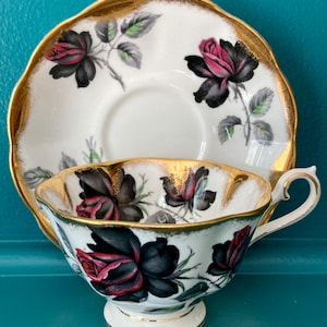 May include: A white teacup and saucer with a black and red rose pattern and gold trim. The teacup and saucer are both decorated with a floral design.
