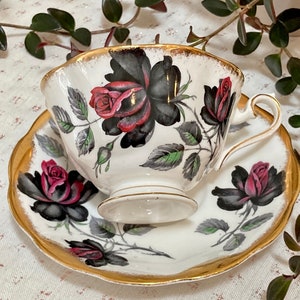 May include: A white teacup and saucer with a black and red rose design. The cup and saucer have a gold trim.
