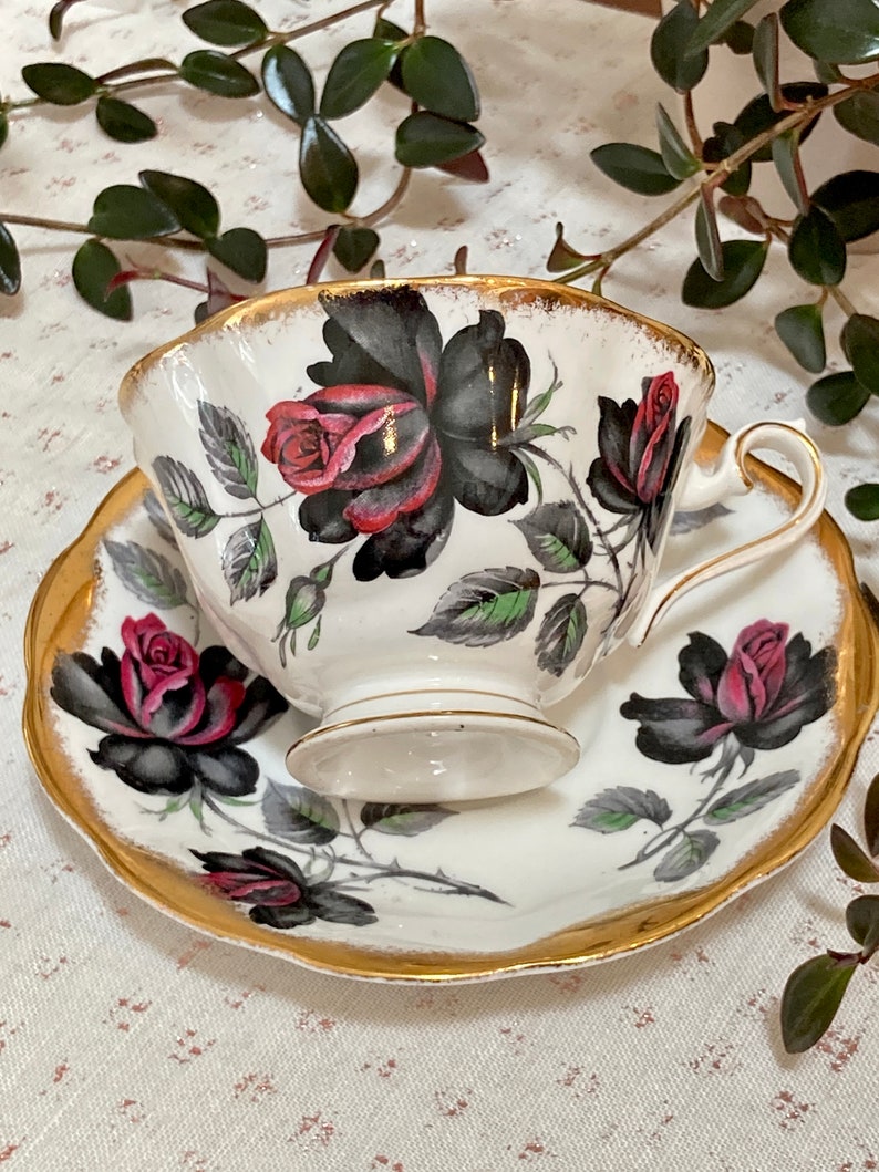 May include: A white teacup and saucer with a black and red rose design. The cup and saucer have a gold trim.