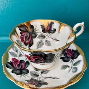May include: A white teacup and saucer set featuring a black and red rose design with gold edging. The set is made of fine china and has a delicate pattern.