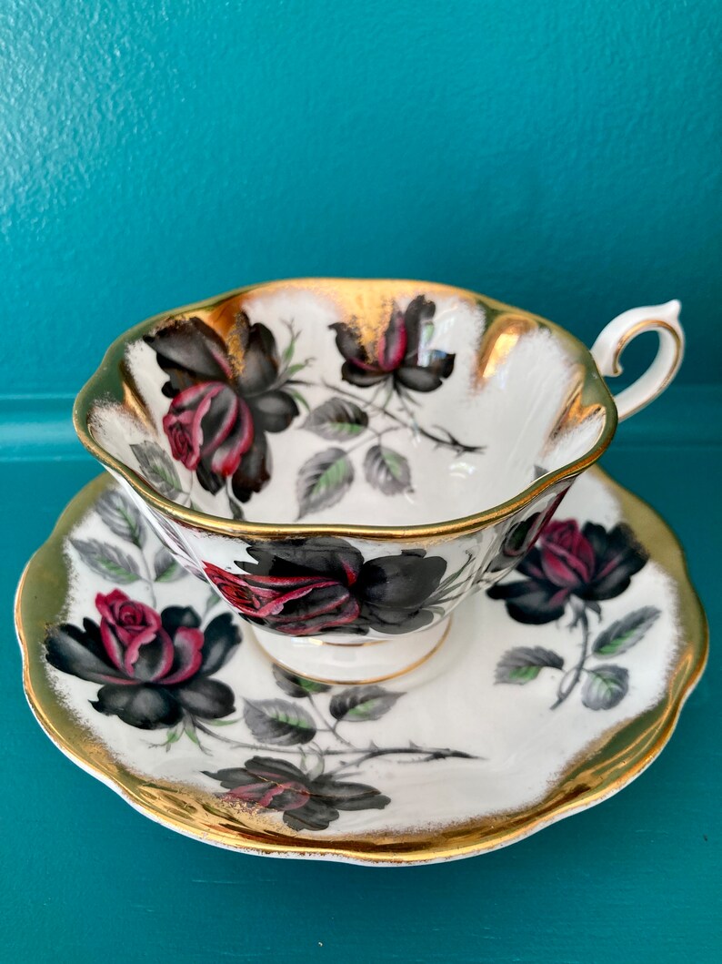May include: A white teacup and saucer set featuring a black and red rose design with gold edging. The set is made of fine china and has a delicate pattern.