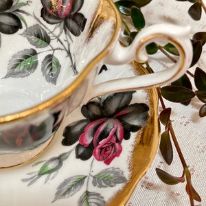 May include: A white teacup and saucer with a gold rim and black and red rose floral pattern.