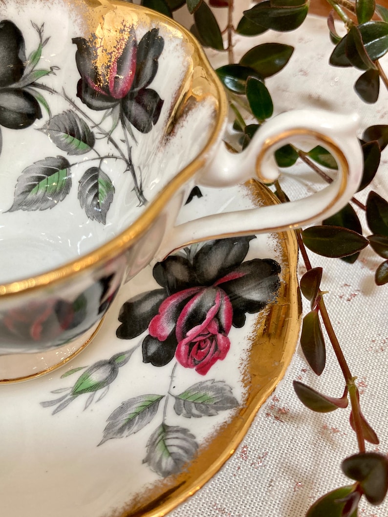 May include: A white teacup and saucer with a gold rim and black and red rose floral pattern.