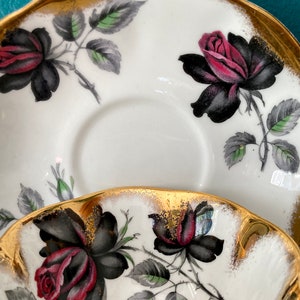 May include: A white teacup and saucer with a gold rim and black and red roses with green leaves.