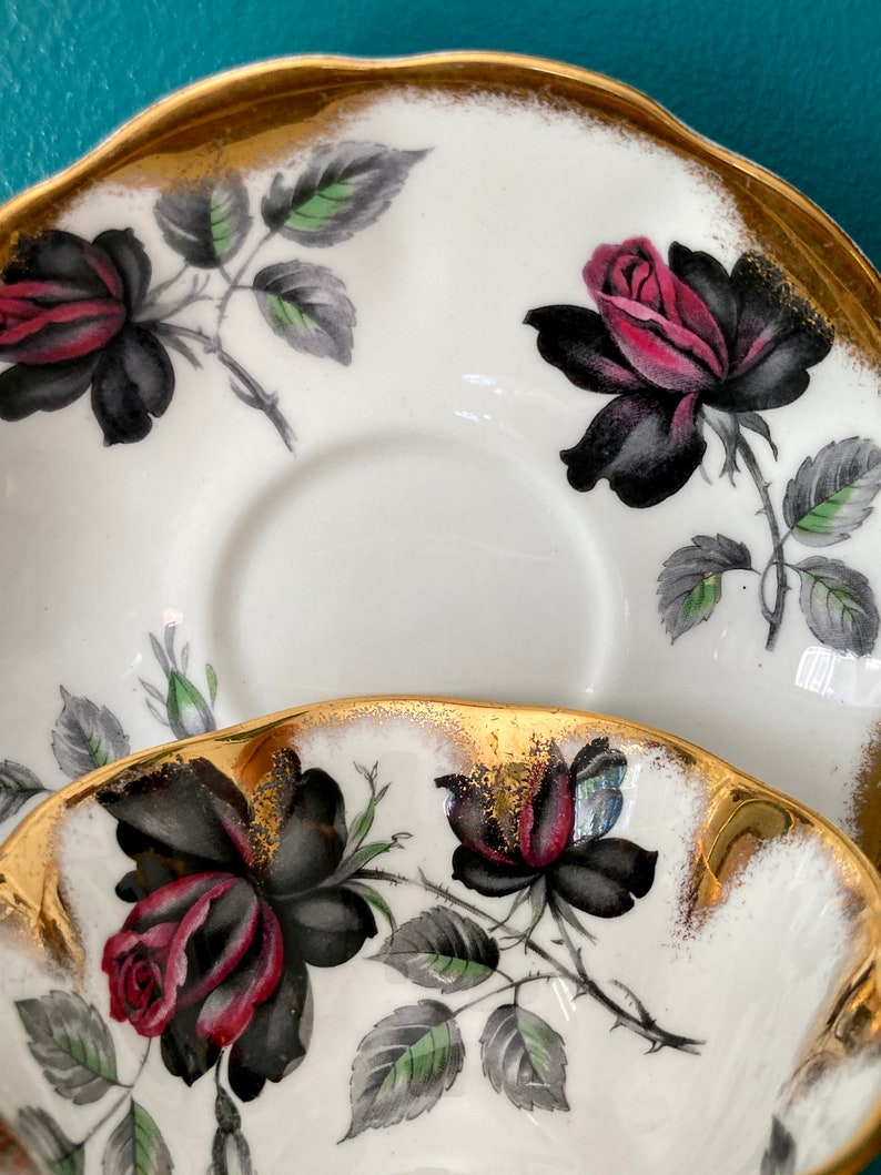 May include: A white teacup and saucer with a gold rim and black and red roses with green leaves.