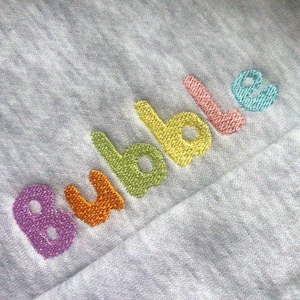 Könnte beinhalten: Nahaufnahme eines hellgrauen Stoffes mit dem gestickten Wort "Bubble". Das Wort ist in einem Regenbogen aus Farben gestickt: Lila, Orange, Grün, Gelb, Rosa und Blau. Der Stoff scheint ein weiches Strickmaterial zu sein.