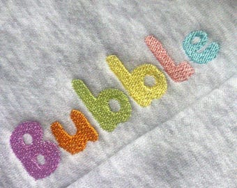 May include: Close-up of a light gray fabric with the embroidered word "Bubble". The word is stitched in a rainbow of colors: purple, orange, green, yellow, pink, and blue. The fabric appears to be a soft knit material.