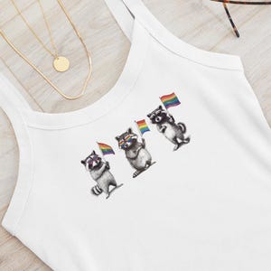 May include: White tank top featuring a graphic of three raccoons holding rainbow pride flags. The top has a scoop neck and wide straps. Sunglasses and a gold necklace are also visible.