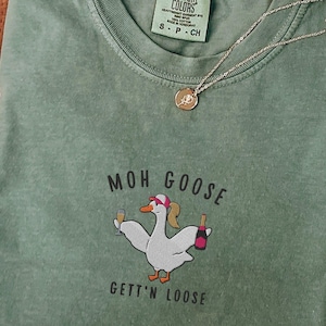 May include: A green t-shirt with the text "Moh Goose Gettin' Loose" and a cartoon goose holding a glass of champagne and a bottle of wine.