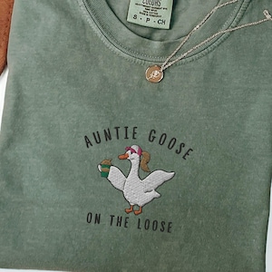 Könnte beinhalten: Ein grünes T-Shirt mit dem Text "Auntie Goose on the Loose" und einem gestickten Bild einer Gans mit einem rosafarbenen Stirnband, die eine Tasse Kaffee hält.