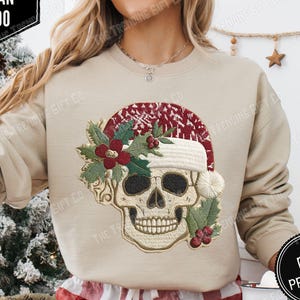 Puede incluir: Sudadera de color arena con una calavera que lleva un gorro de Papá Noel adornado con acebo y bayas. El gorro tiene un estampado rojo y blanco. La sudadera tiene el texto "DTG PRINTED" en un círculo negro. La marca es Gildan 18000.