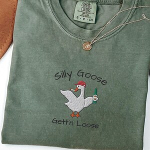 May include: A green t-shirt with an embroidered image of a goose wearing a red hat and holding a beer bottle. The text "Silly Goose Gettin' Loose" is embroidered below the image.