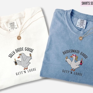 May include: Two white t-shirts with a goose graphic and the text "Silly Bride Goose Gett'n Loose" and "Bridesmaid Goose Gett'n Loose". The shirts are on a white background. The shirt on the left is ivory and the shirt on the right is blue jean.