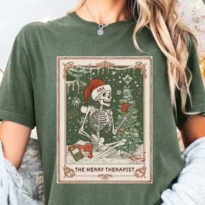 May include: Moss green t-shirt featuring a graphic of a skeleton wearing a Santa hat, holding a mug, and sitting by a Christmas tree. The graphic is framed with the text "THE MERRY THERAPIST".