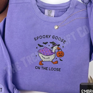 Peut inclure: Sweat-shirt violet Comfort Colors avec un motif brodé d'Halloween. Le motif représente une oie portant un chapeau de sorcière et une citrouille, avec le texte "Spooky Goose On The Loose".