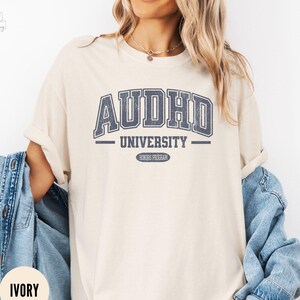 May include: A beige t-shirt with a blue graphic that reads "AUDHD University - Honours Program".