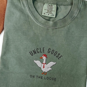 May include: A moss green t-shirt with the text "Uncle Goose On The Loose" and an embroidered image of a goose wearing a red hat and a white scarf.