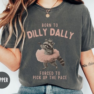May include: A gray t-shirt with the text "Born to Dilly Dally" and "Forced to Pick Up the Pace" in pink. A cartoon raccoon wearing a pink tutu and a crown is in the center of the shirt.