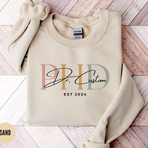 Könnte beinhalten: Ein sandfarbener Pullover mit einem grafischen Design, das "PhD Dr. Custom EST 2024" in einer stilisierten Schrift mit rosa, goldenen und grünen Buchstaben zeigt.