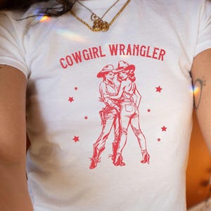 Könnte beinhalten: Weißes T-Shirt mit dem roten Schriftzug "COWGIRL WRANGLER" und einer roten Grafik von zwei Frauen in Cowboyhüten und -stiefeln. Rote Sterne sind um die Grafik herum verteilt. Das T-Shirt ist aus weichem Material.