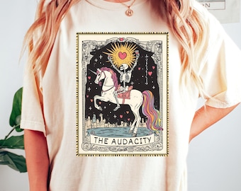 May include: A white t-shirt featuring a graphic of a skeleton riding a rainbow unicorn. The graphic is in a tarot card style with the text "THE AUDACITY".