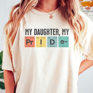 Pode incluir: Camiseta de cor marfim com o texto "My Daughter, My Pride" em letras pretas. O texto é estilizado para parecer uma tabela periódica com as letras "Pr", "I", "D" e "e" cada uma em uma caixa de cor diferente com o número atômico e o símbolo do elemento correspondente.