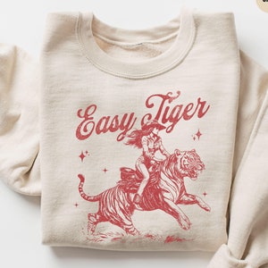 May include: A beige sweatshirt with a red graphic print of a woman riding a tiger. The graphic includes the text "Easy Tiger".