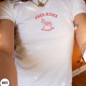 May include: White t-shirt with the text "FREE RIDES" above a red rocking horse graphic. The shirt has a crew neck and short sleeves. The design is centered on the chest.