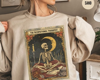 Może przedstawiać: Beżowa bluza z vintage'ową ilustracją szkieletu siedzącego w medytacyjnej pozie pod rosnącym księżycem. Ilustracja jest otoczona ozdobną ramką i zawiera tekst "The Occupational Therapist".