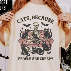May include: Ivory t-shirt with a graphic of a skeleton on a sofa with black cats and bats. The text says "CATS, BECAUSE PEOPLE ARE CREEPY." The shirt has a round neck and short sleeves.
