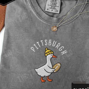 Op de afbeelding: Pepperkleurig t-shirt met een geborduurd ontwerp van een gans met een gele hoed en een voetbal. Het woord "Pittsburgh" is boven de gans gebogen. Het shirt heeft een Comfort Colors label.
